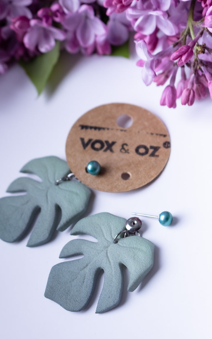 Boucles d’oreilles MAUI Lagoon édition limitée avec feuilles Monstera vert lagon et perles d’eau douce turquoise présentées sur carte Vox &amp; Oz avec fleurs lilas