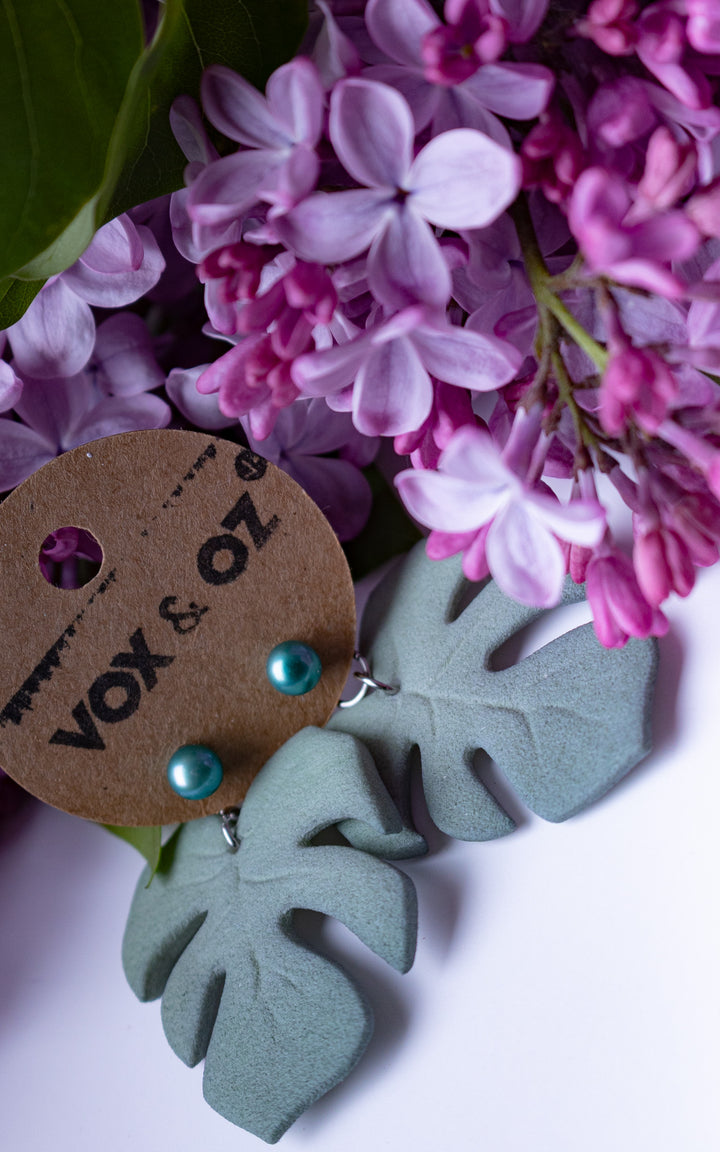 Boucles d’oreilles MAUI Lagoon édition limitée avec feuilles Monstera vert lagon et perles turquoise présentées avec fleurs lilas et carte Vox &amp; Oz